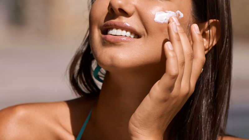 Ilustrasi seorang perempuan sedang mengoleskan sunscreen di wajahnya agar kulitnya tidak terbakar sinar matahari.
