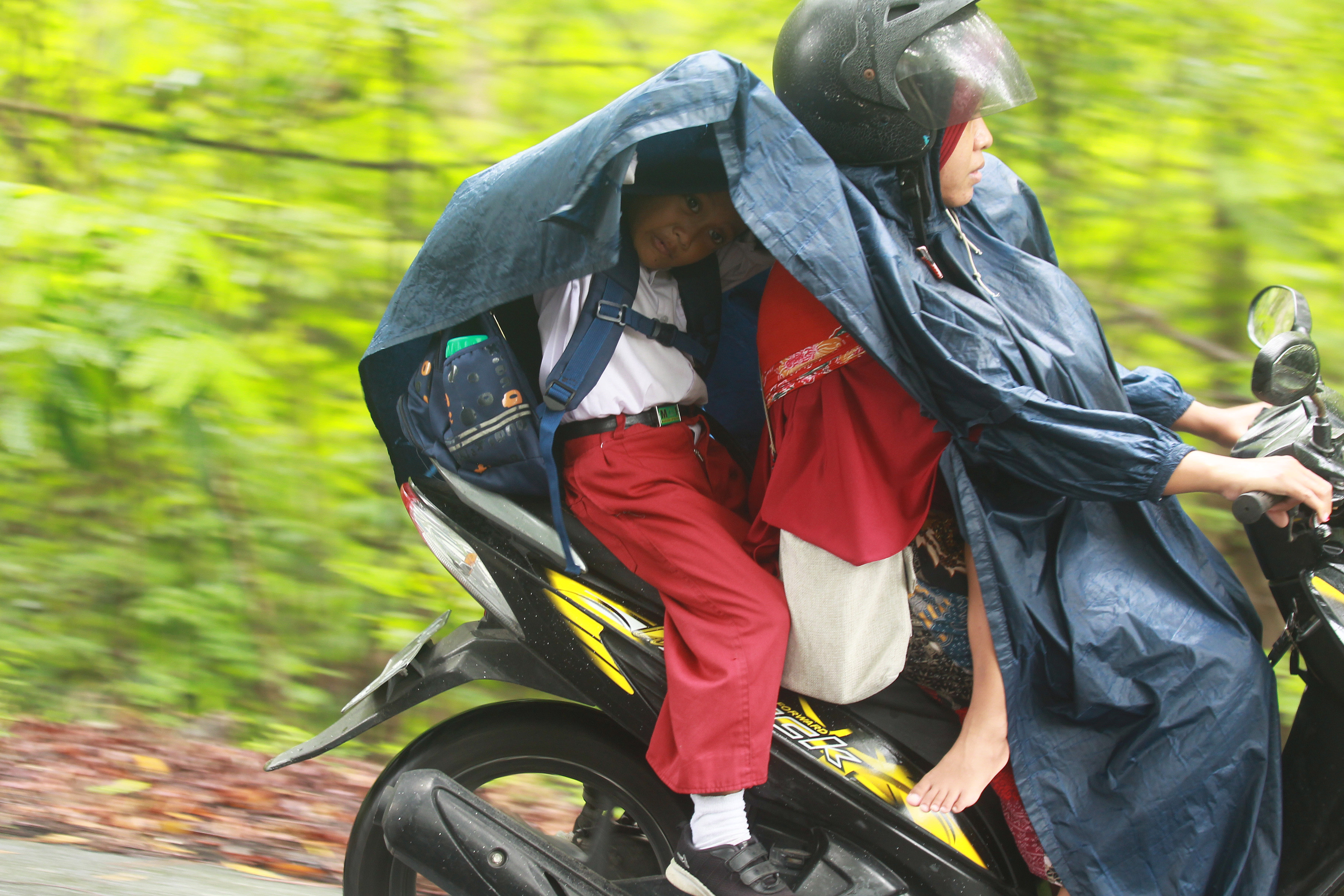 Seorang siswa sekolah berlindung dari air hujan dengan mantel saat diantar ibunya dengan motor