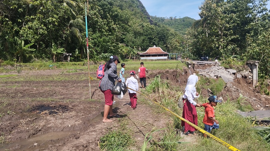 HARAP PERBAIKAN: Wali murid SDN Kadung Miri sedang menyeberangkan anaknya setelah pulang sekolah melewati Jalan di Wunur yang Ambrol&nbsp;Senin (23/11/2025).