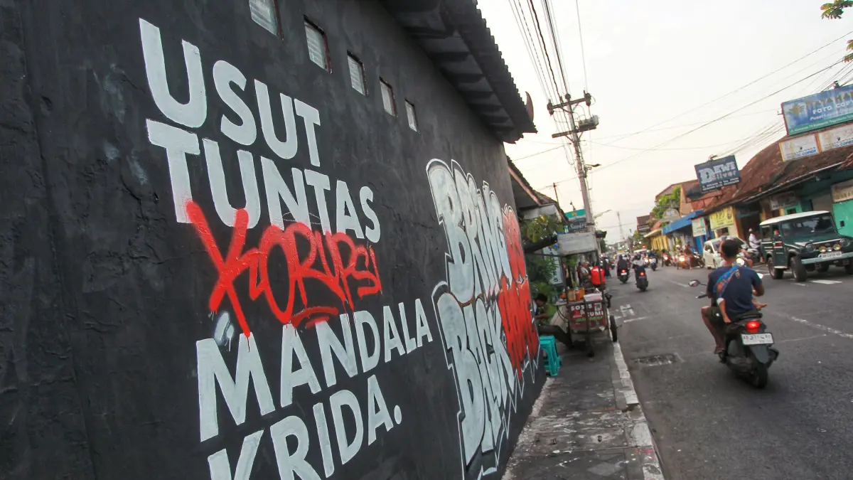 Pengguna jalan melintas di dekat mural bertuliskan "Usut Tuntas Korupsi Mandala Krida. Bring Us Back Home", di Jalan Kemasan, kawasan Kotagede, Kota Jogja, kemarin (1/6)