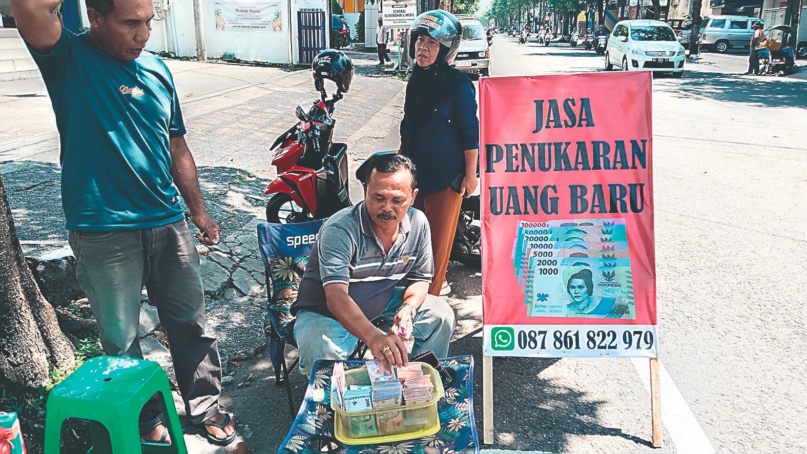 Seorang pedagang jasa penukaran uang baru melayani pelanggan di pinggir jalan.