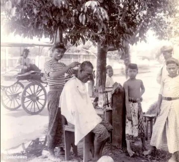 Tukang potong rambut asal Madura sedang memangkas rambut pelanggannya di Surabaya tahun 1930-an (KITLV).