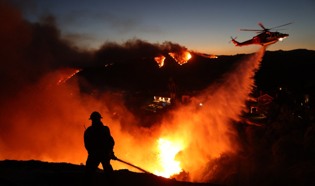 Personel pemadam kebakaran menghadapi rumah-rumah yang hancur saat helikopter melepaskan air saat Palisades Fire membesar di Pacific Palisades. Kebakaran ini belum pernah terkendali sama sekali