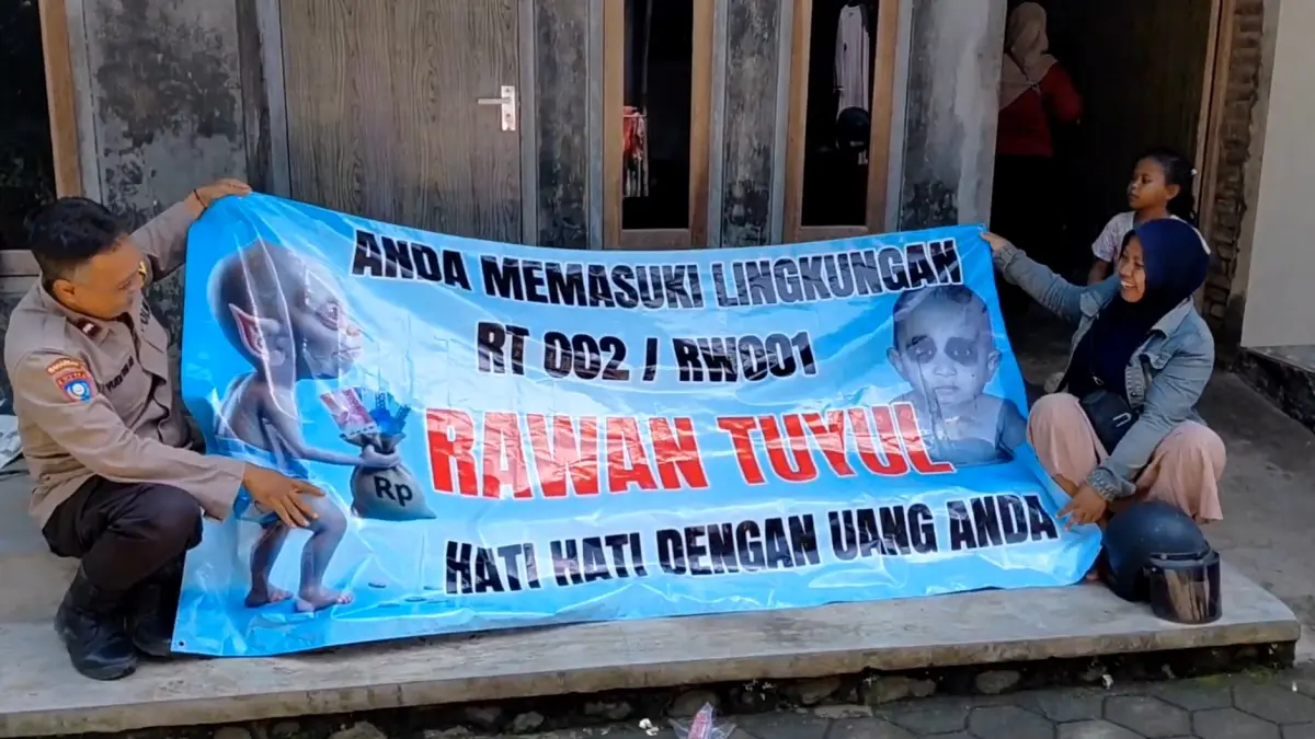 BIKIN HEBOH: Banner biru bergambar tuyul “nongol” di pintu masuk Dusun Mencek, Desa Serut, Kecamatan Panti. (M ADHI SURYA/RADAR JEMBER)