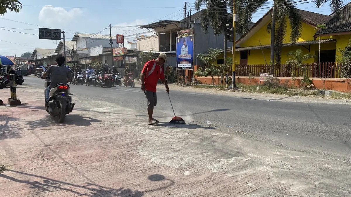 PENJAGA KESEIMBANGAN JALAN: Sahid, PHL Polsek Wuluhan, dengan telaten menyapu ceceran pasir di Jalan Pahlawan, Desa Dukuhdempok, demi memastikan pengendara motor terbebas dari ancaman terpeleset. (JUMAI/RADAR JEMBER)