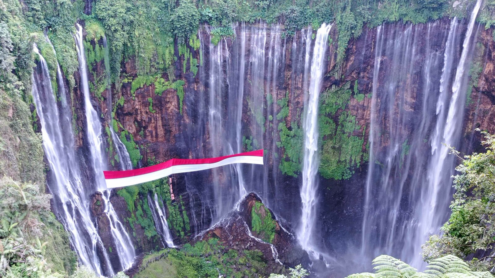 MERAH PUTIH: Bendera merah putih dengan panjang 150 meter dan lebar enam meter terbentang di antara Air Terjun Tumpak Sewu. Bentangan bendera dilaksanakan untuk memperingati HUT Polri ke-75 tahun.