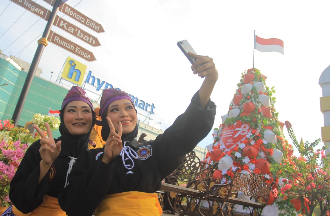 UNIK: Dua remaja putri berswafoto di depan tumpeng raksasa yang dibebat caping dan hasil bumi di kawasan Pahlawan Religi Center kemarin (12/8).&nbsp;(BAGAS BIMANTARA/RADAR MADIUN)