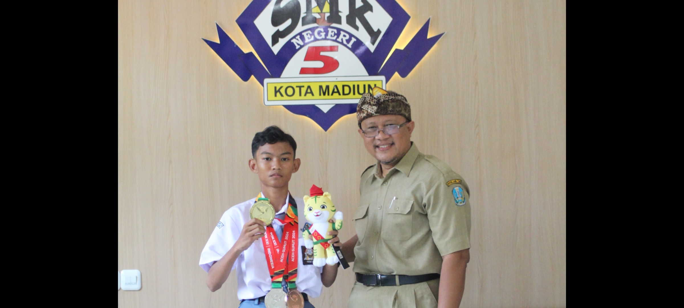 BANGGA: Kepala SMKN 5 Kota Madiun Djuniedi Ekosetiono bersama Surya Adi Putra menunjukkan medali emas dan maskot PON 2024. (ABRIANTO WIBOWO/RADAR MADIUN)