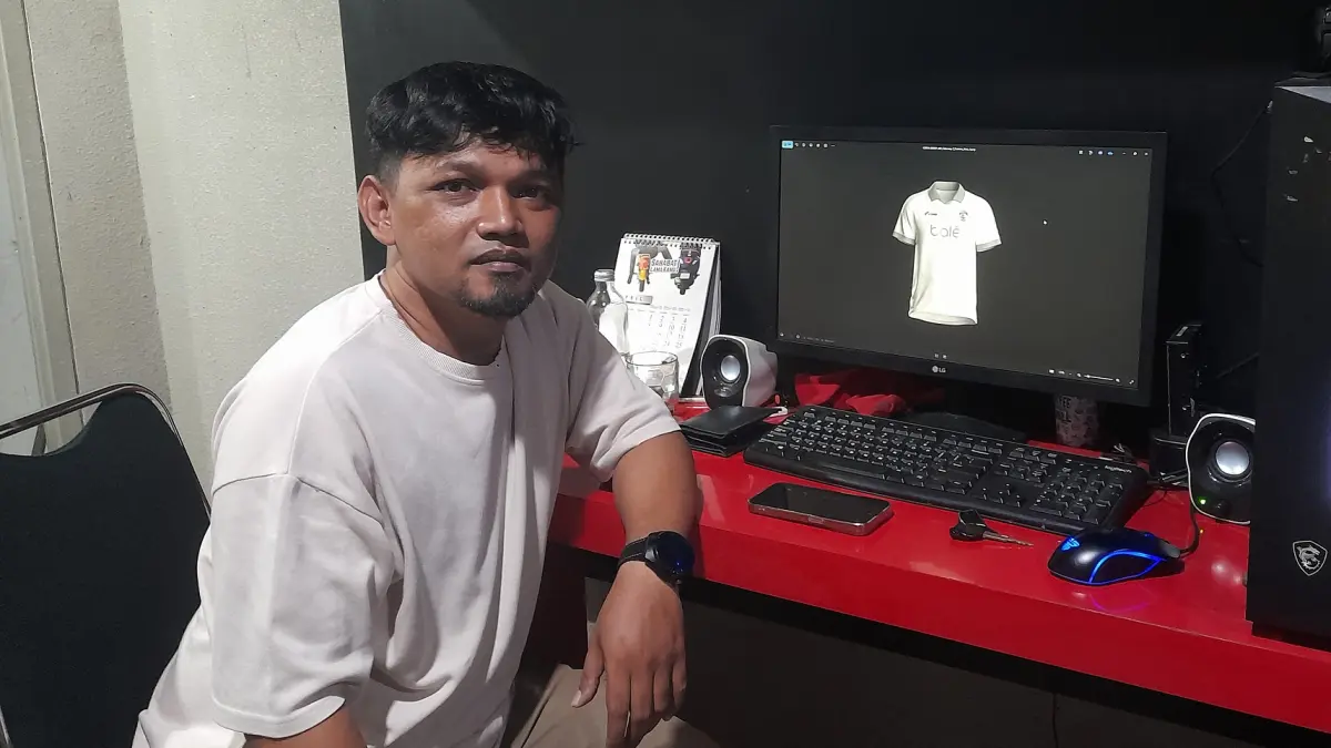 PENUH DEDIKASI: Heni Kurniawan foto dengan berlatar belakang desain jersey keempat Arema FC yang dibuatnya kemarin siang. Dia mendesain empat jersey yang digunakan pengawa Singo Edan musim 2025/2026. (MA Fauzan/Radar Malang)