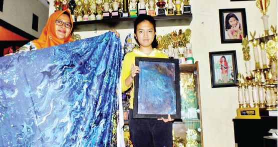 Athaya (kanan) bersama Maisah, ibunya, menunjukkan lukisan yang juga dicetak di atas kain. Di belakang mereka tampak deretan piala lomba modeling yang diraih Athaya semasa kecil. (Achmad Fikyansyah/Radar Malang)