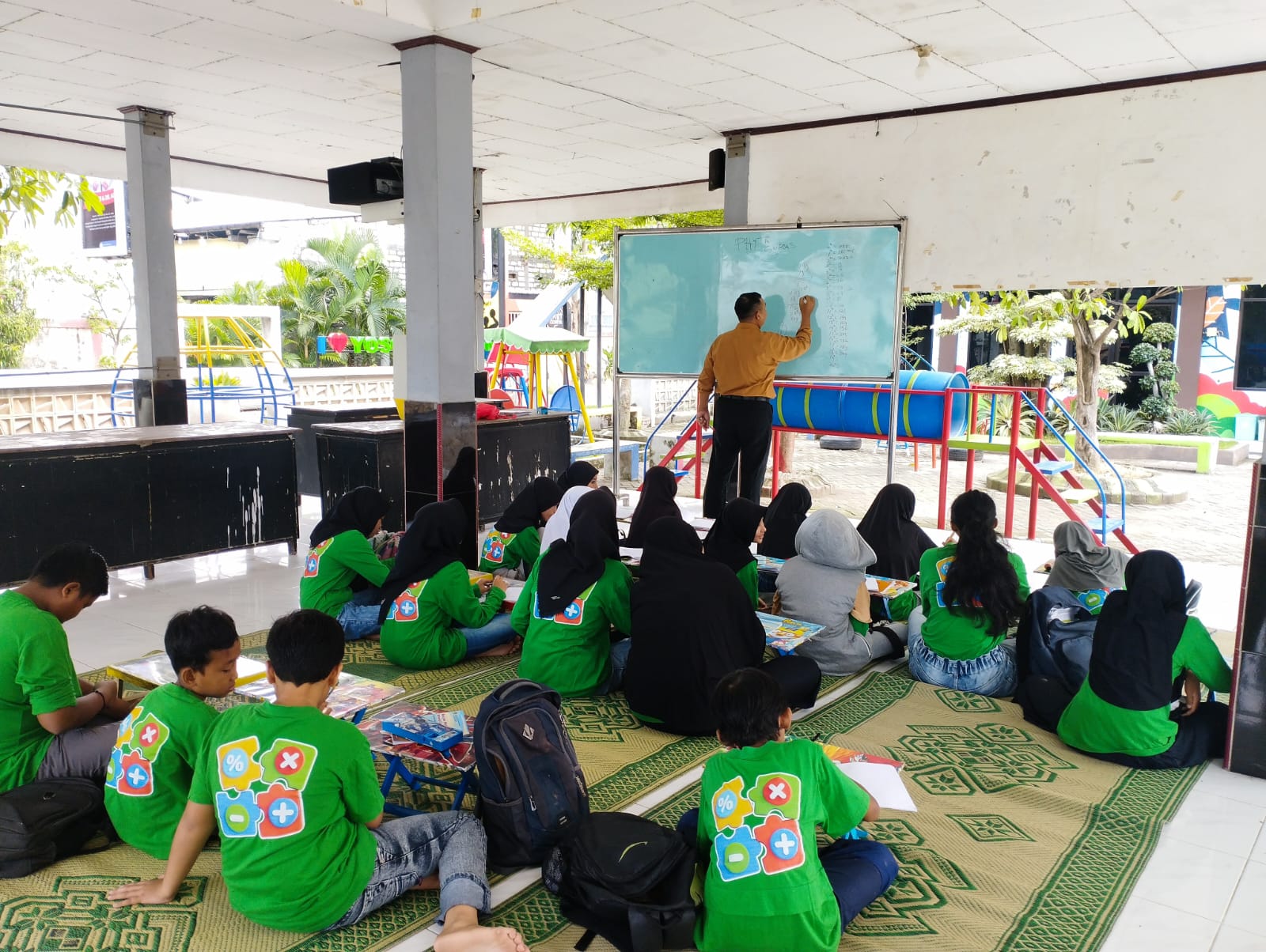 PRODUKTIF: Puluhan anak-anak tampak duduk rapi di atas tikar, fokus menyimak materi pelajaran matematika yang disampaikan oleh pengajar di papan tulis.