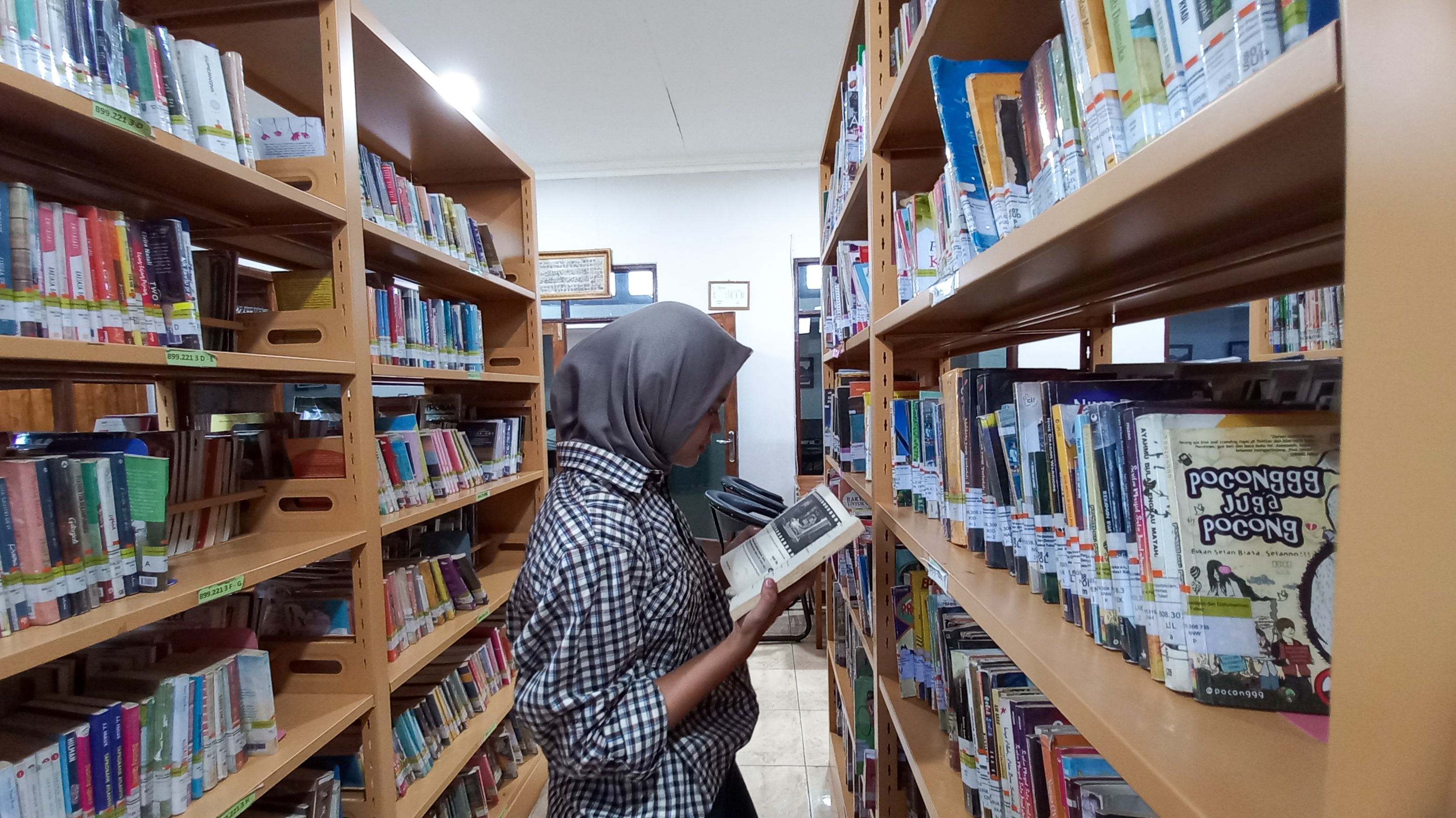 Salah seorang pengunjung di Perpustakaan Daerah Tuban sedang membaca novel di antara rak buku kesusastraan.