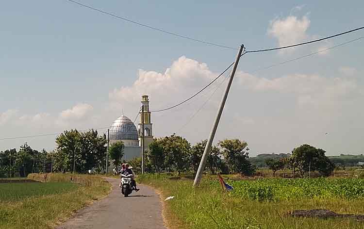RAWAN ROBOH: Pengendara motor melintas di sekitar tiang listrik PLN yang miring ke selatan di Desa Randuputih, Kecamatan Dringu.