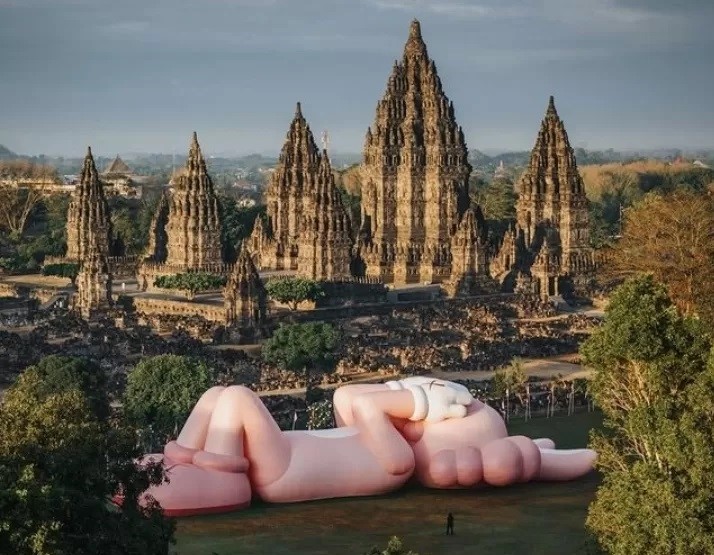 Boneka raksasa KAWS rebahan di pelataran Candi Prambanan.
