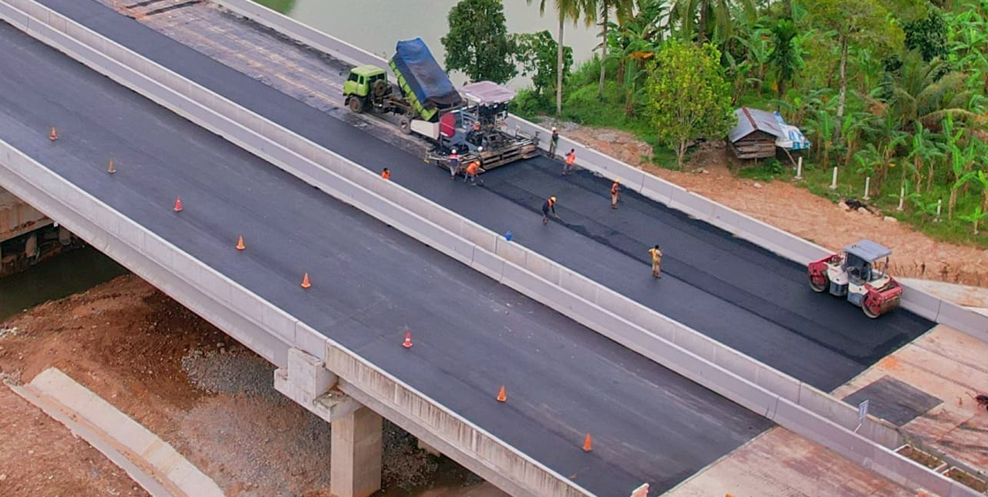 Pekerja dan alat berat bekerja melapisi aspal hotmix pada permukaan atas jembatan jalan tol.