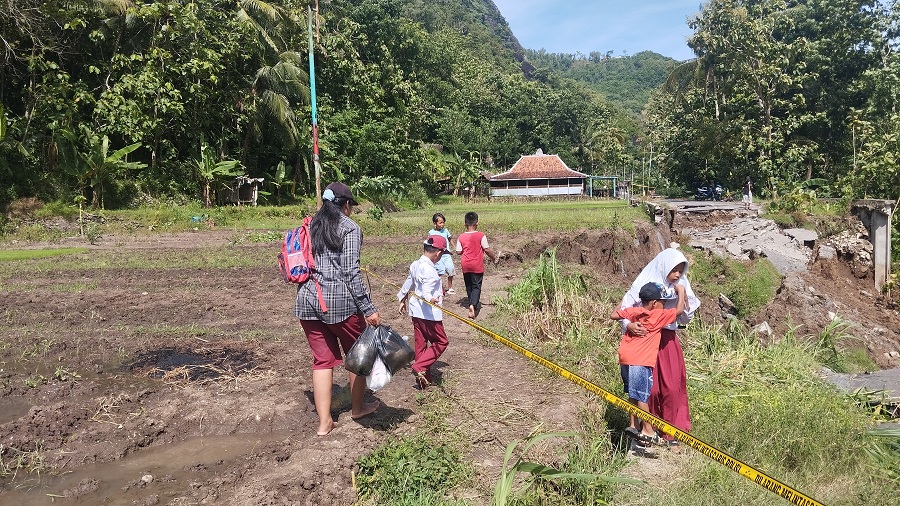 HARAP PERBAIKAN: Wali murid SDN Kadung Miri sedang menyeberangkan anaknya setelah pulang sekolah melewati Jalan di Wunur yang Ambrol&nbsp;Senin (23/11/2025).