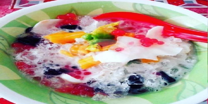 Semangkuk es campur warna-warni dengan berbagai buah segar, mutiara merah, dan serutan kelapa muda.