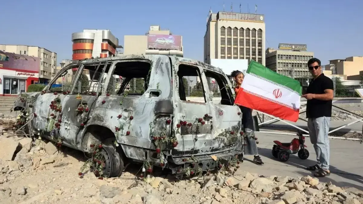 Seorang pria mengibarkan bendera Iran di samping ambulans Bulan Sabit Merah Iran yang hancur akibat serangan udara Israel. (Al Jazeera)

