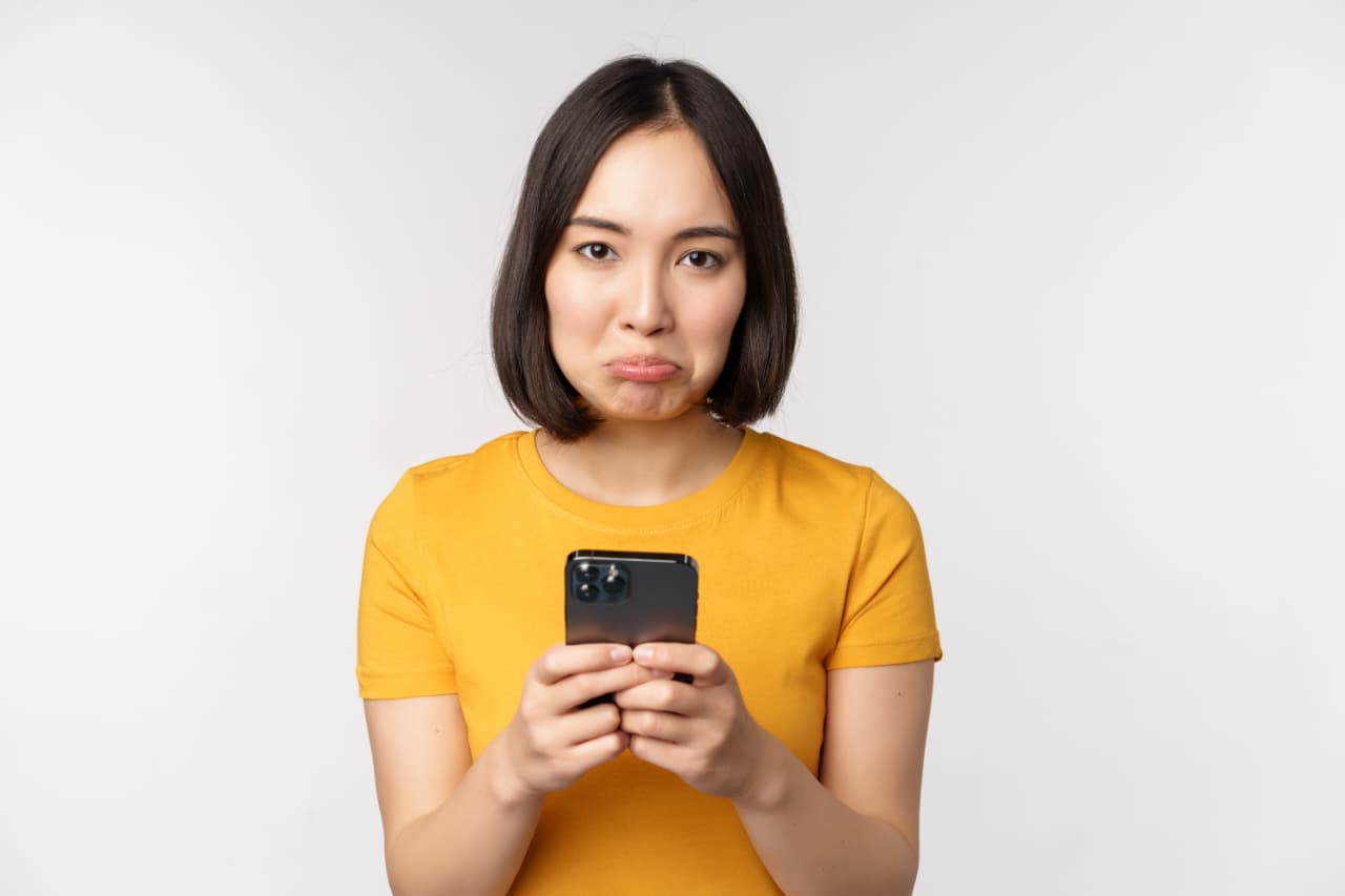 Seorang wanita yang sedang memegang ponsel dengan raut wajah cemberut