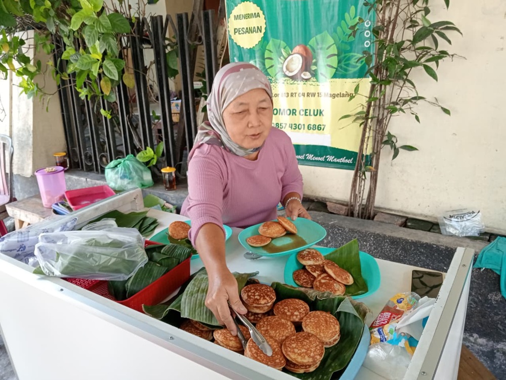 Indah sedang menyusun apem yang baru matang di atas nampan beralas daun pisang.