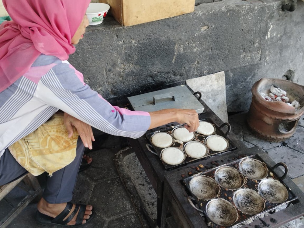Tini sedang memanggang apem di atas panggangan besi