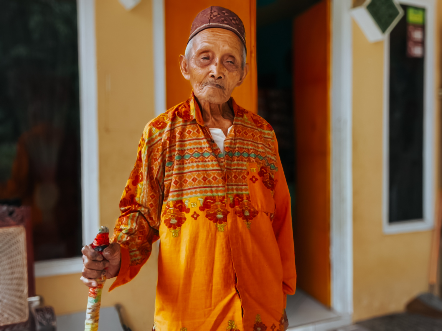 Mbah Sastro berdiri di depan rumahnya sambil memegang tongkat kesayangan. Meski berusia 103 tahun, ia masih terlihat bugar dan tetap menjalankan aktivitas ibadah setiap hari.