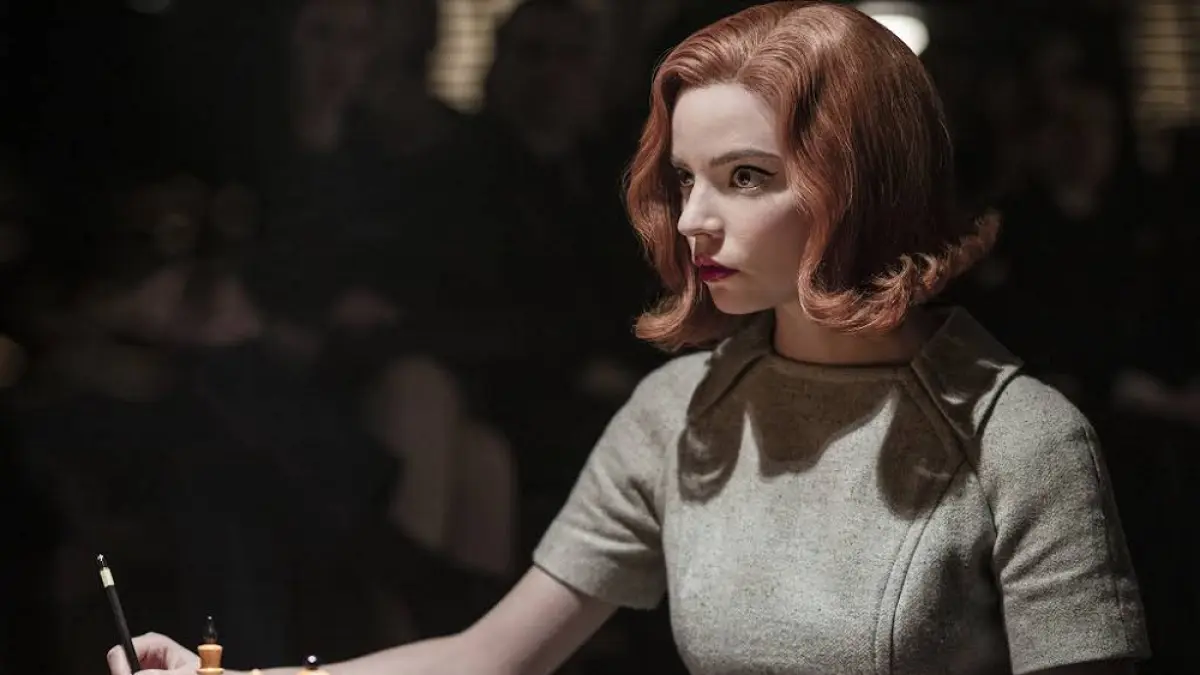 Beth Harmon (Anya Taylor-Joy) tampak fokus menatap papan catur sambil memegang bidak putih, dengan pencahayaan dramatis khas adegan turnamen di The Queen