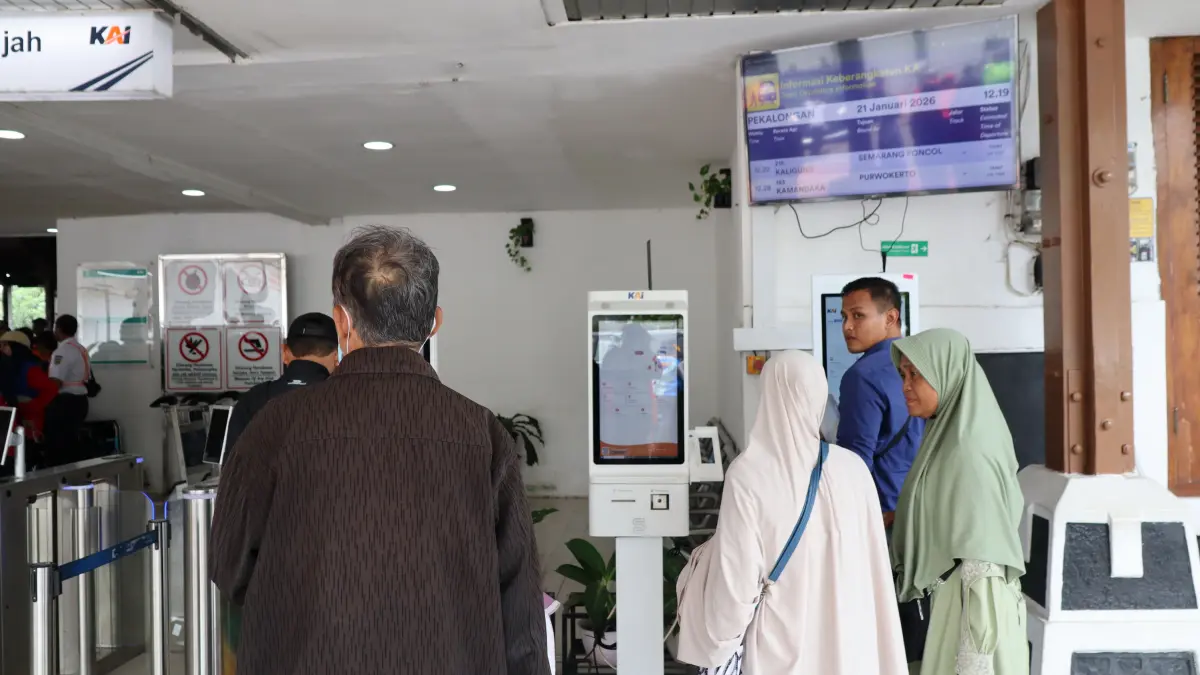 Commuters line up at self-service ticket machines inside a busy train station, printing tickets while others check schedules on a digital departure board above, Pekalongan, Indonesia, January 21, 2026