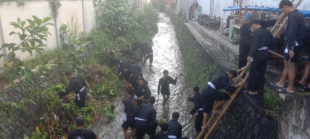 PEDULI LINGKUNGAN: Anggota PSHT Kota Blitar membersihkan aliran sungai dari sampah dan rumput liar, Sabtu (14/6/2025).