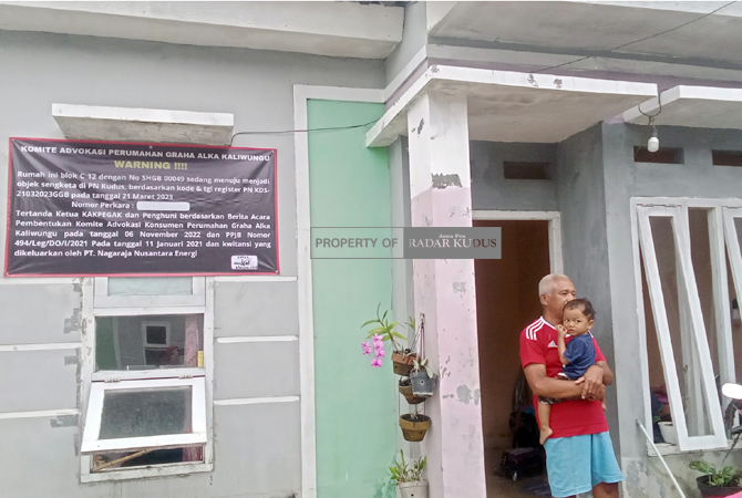 DIPASANG BANNER: Susanto, penghuni rumah di Perumahan Graha Alka Blok C 12 bersama cucunya kemarin. (ARIKA KHOIRIYA/RADAR KUDUS)