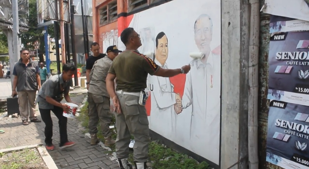 BERSIHKAN: Petugas Satpol PP dan Bawaslu Blora membersihkan APK berbentuk mural dengan cat