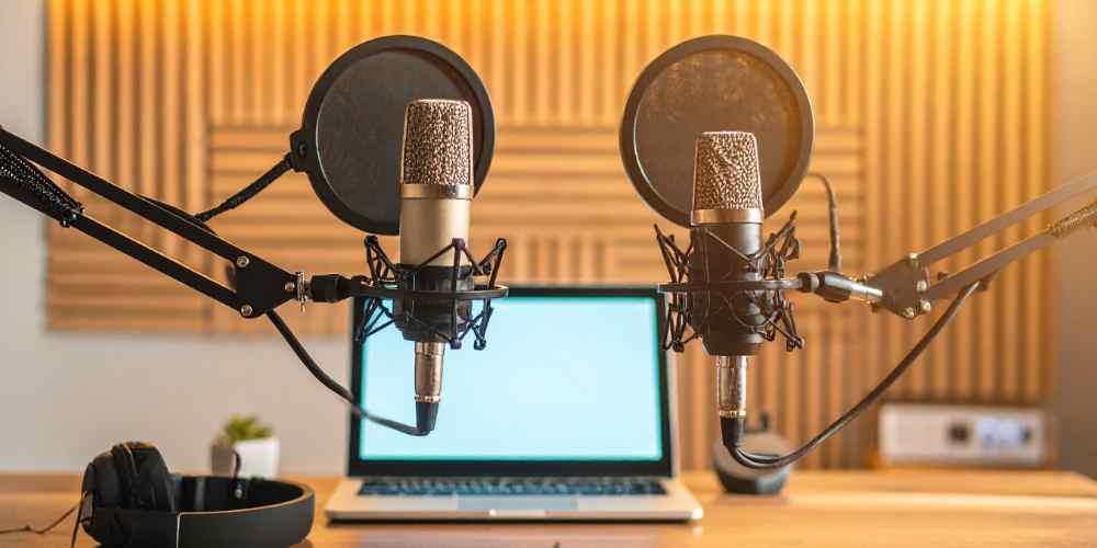 Dua mikrofon studio dengan pop filter menghadap ke sebuah laptop, menggambarkan suasana ruang rekaman podcast yang profesional dan nyaman.