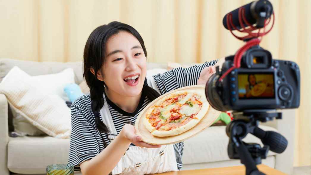 Seorang food vlogger sedang merekam konten sambil memperlihatkan pizza yang baru dibuat, menunjukkan antusiasme dan keceriaan khas video kuliner yang menarik perhatian penonton.