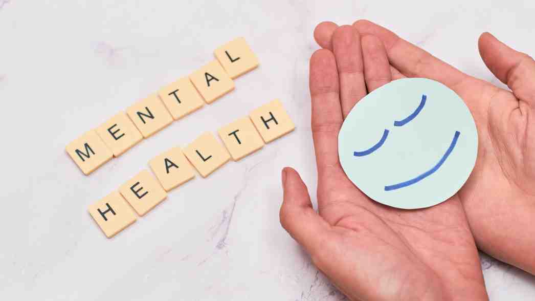 Ilustrasi kesehatan mental: simbol wajah tenang di atas telapak tangan dan huruf scrabble bertuliskan &ldquo;MENTAL HEALTH&rdquo;, menggambarkan pentingnya menjaga ketenangan diri dan kesejahteraan emosional.