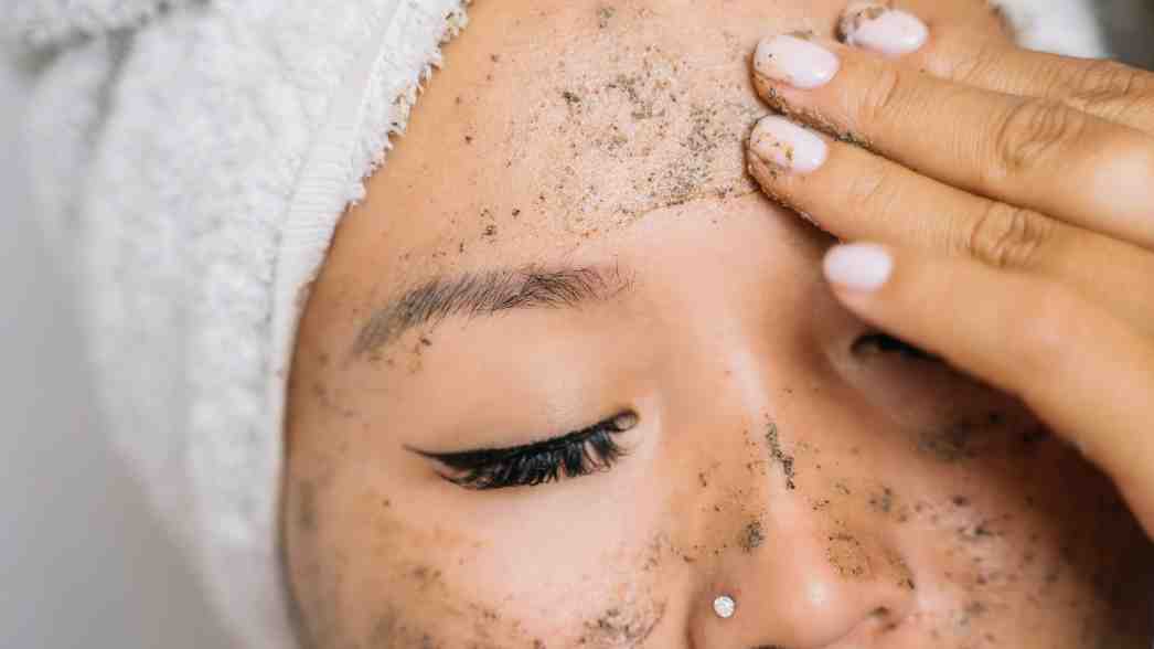Seorang perempuan sedang mengaplikasikan scrub wajah, memperlihatkan proses eksfoliasi untuk mengangkat sel kulit mati dan membuat kulit tampak lebih halus.