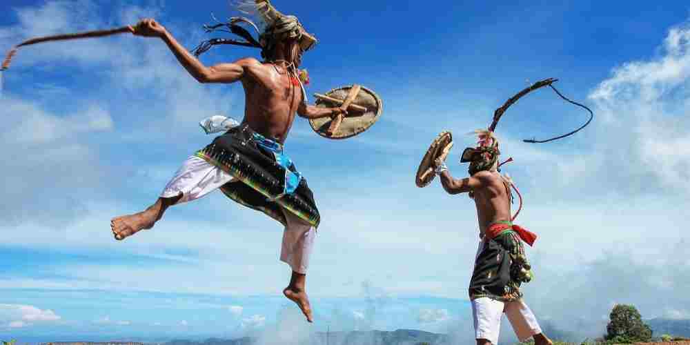 Dua penari Tari Caci saling beradu ketangkasan di udara, memvisualkan keberanian, sportivitas, dan kehormatan dalam tradisi adat Manggarai, Flores, Nusa Tenggara Timur.