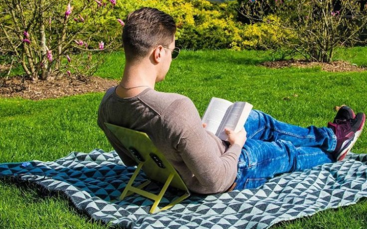 Seseorang sedang berjemur sambil membaca buku di rerumputan taman.