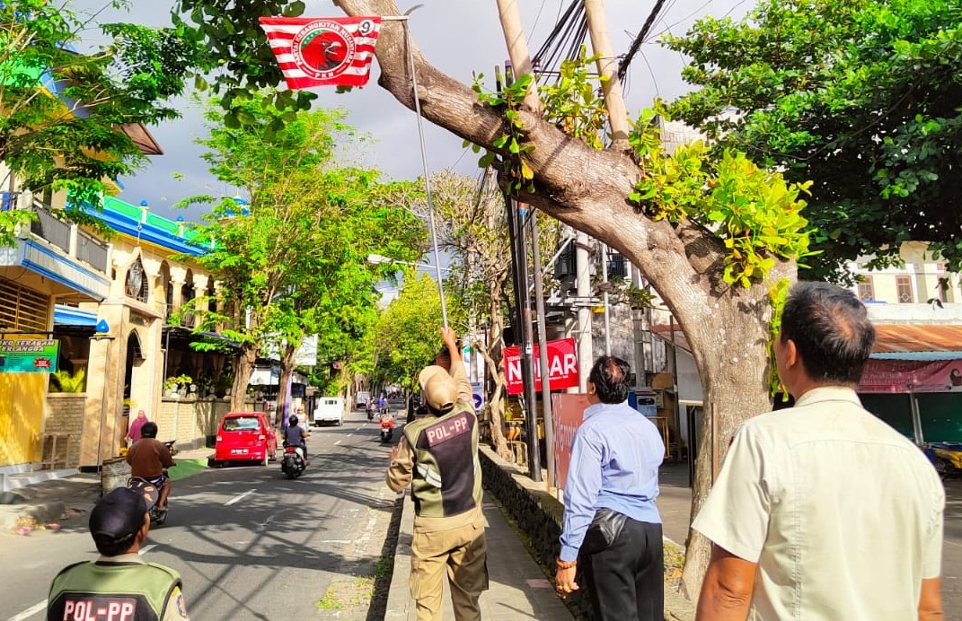 DITURUNKAN: Petugas Satpol PP Karangasem saat menurunkan atribut partai, pamflet hingga baliho yang terpasang di pohon perindang seputaran kota Amlapura, Kamis (20/7/2023).