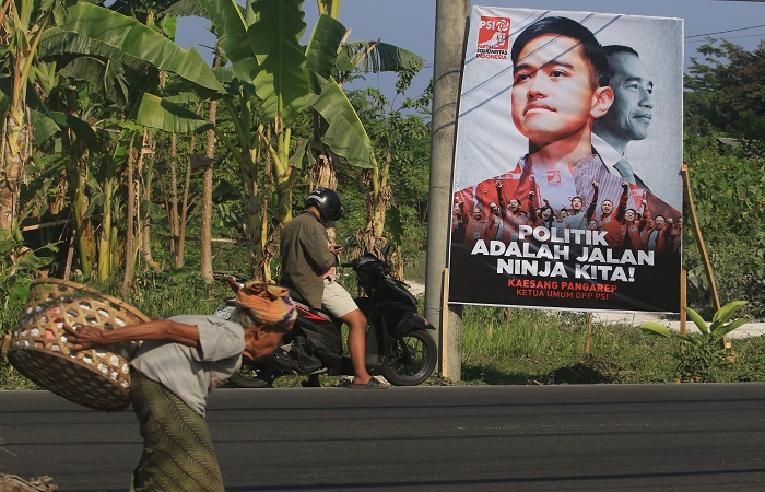 Seorang nenek sambil memikul beban melewati baliho putra bungsu presiden Jokowi, Kaesang Pengarep di Jalan Hayamwuruk Denpasar.
