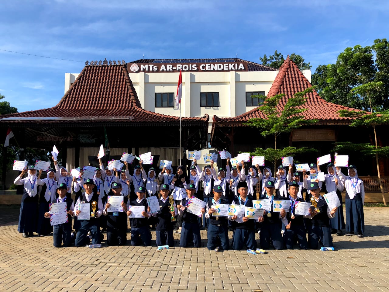Siswa-siswi MTs Ar-Rois Cendekia berfoto bersama dengan trofi dan medali yang diterima dari perlombaan, di halaman MTs Ar-Rois Cendekia.
