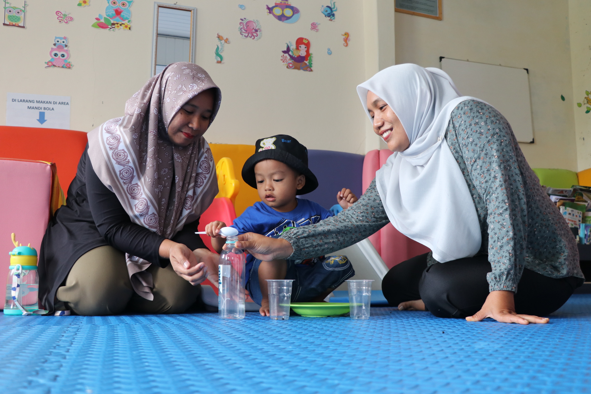 Fasilitator Rumah Anak SIGAP Sokawera Parsini (kanan) sedang belajar motorik halus pada anak dengan memasukkan biji kedelai bersama orang tua.