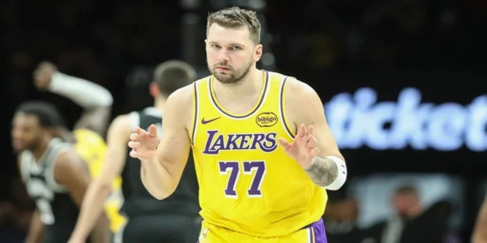 Luka Doncic, bintang Los Angeles Lakers, terlihat mengenakan jersey kuning bernomor 77 saat berada di lapangan. Ia mengangkat kedua tangannya dengan ekspresi fokus, seolah memberi arahan atau merespon