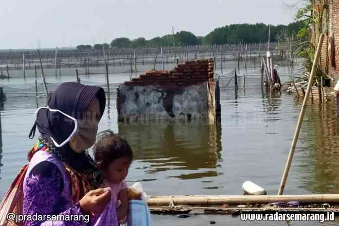 Seorang ibu menggendong anaknya melalui jalan Desa Bedono, Kecamatan Sayung yang sekitarnya tampak bangunan rumah dan tambak hancur dikepung rob. (Wahib Pribadi/Jawa Pos Radar Semarang)