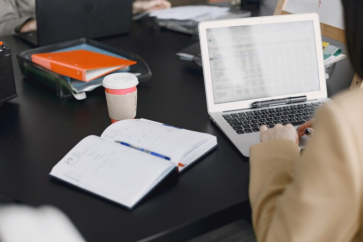 FOKUS: Seorang warga sedang mengetik di laptop dikelilingi buku catatan, bolpoin, dan cangkir kopi di meja. (FREEPIK UNTUK RADARMADURA.ID)