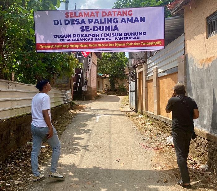 PROTES: Warga berjalan di dekat spanduk bertulis sindiran yang dipasang di Dusun Pokapoh, Desa Larangan Badung, Kecamatan Palengaan, Pamekasan, Kamis (21/8). (MOH. IQBAL AFGANI/JPRM)