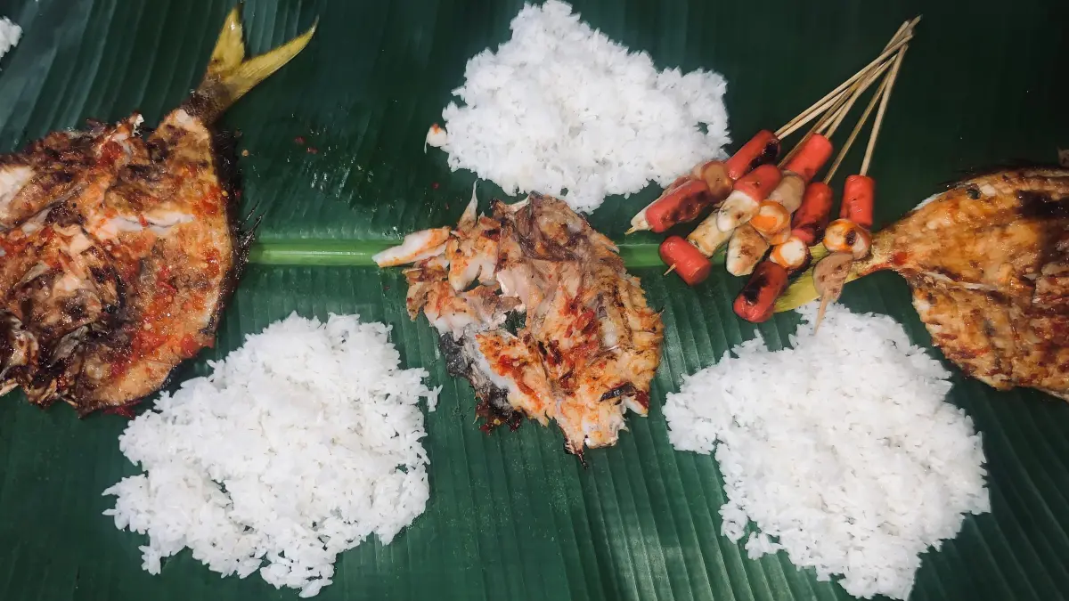 Ikan bakar hangat disajikan di atas daun pisang, lengkap dengan nasi, sambal, dan lalapan. Penyajian sederhana di tempat lesehan menambah kesan santai saat bersantap bersama keluarga maupun teman. (Desi untuk Radar Banyuwangi)